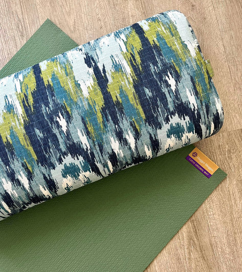 Hugger Mugger Yoga Bolster in Blue Lagoon pattern rests on a green yoga mat, ideal for enhancing yoga practice. The bolster features a vibrant, abstract design with shades of blue, green, and white. Perfect for providing support and comfort during